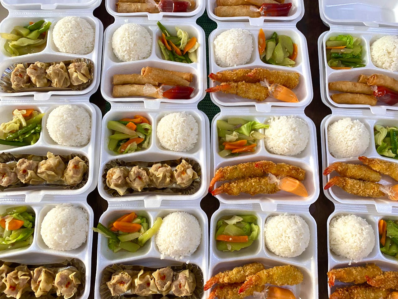 Top view of various Asian meals with rice, vegetables, and dumplings in take-out containers.