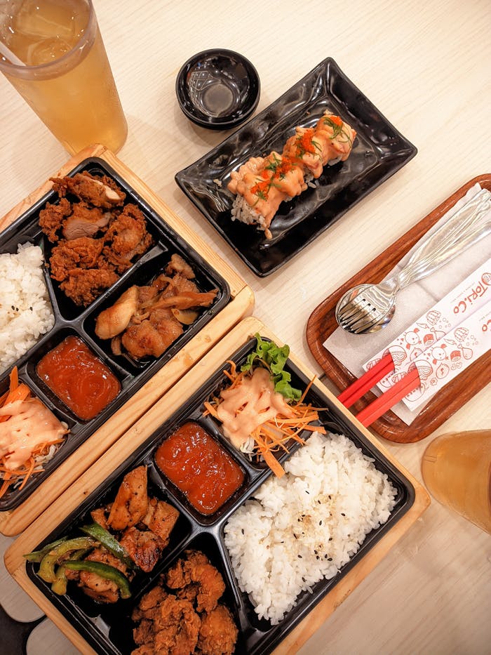 A top-down view of a Japanese bento box meal with sushi, drinks, and chopsticks on a wooden table.