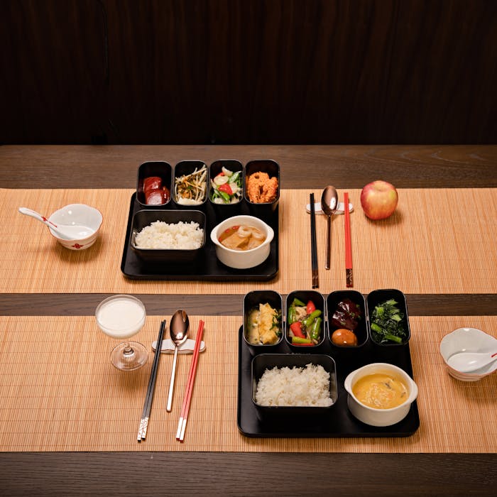 journey Two Japanese bento box meals with rice, vegetables, and soup on a bamboo table setting.